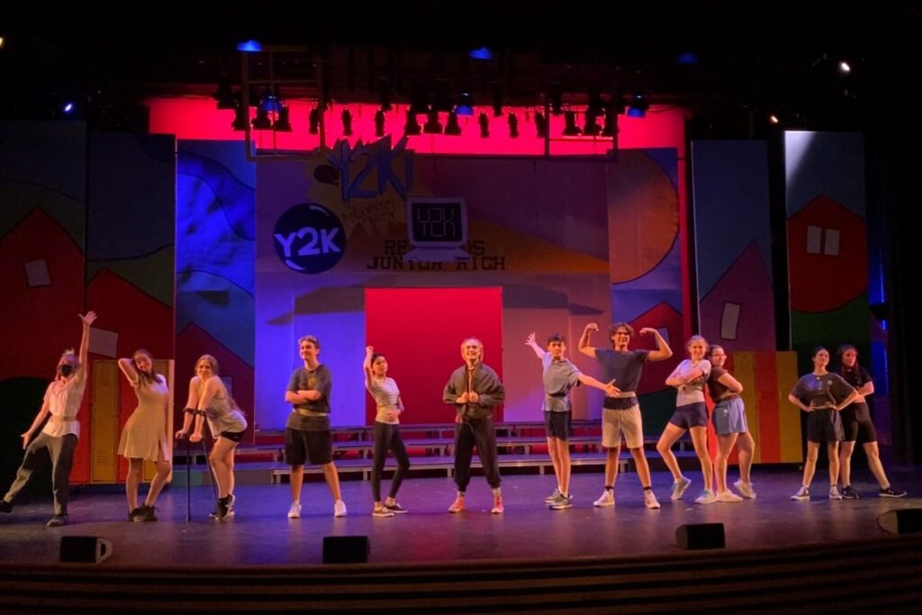 Students from the Kay Meek Youth Conservatory are posing on a dark stage with a colourful backdrop