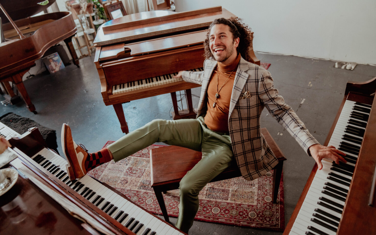 Emmet Cohen leans back on a piano stool smiling with his hands up on two pianos and his foot up on another